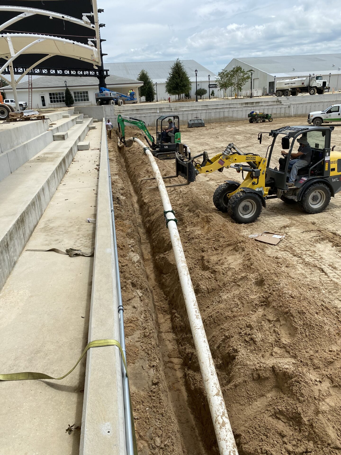 Construction site with machinery and workers installing a long pipe in a trench beside concrete seating steps, under a large metal canopy.