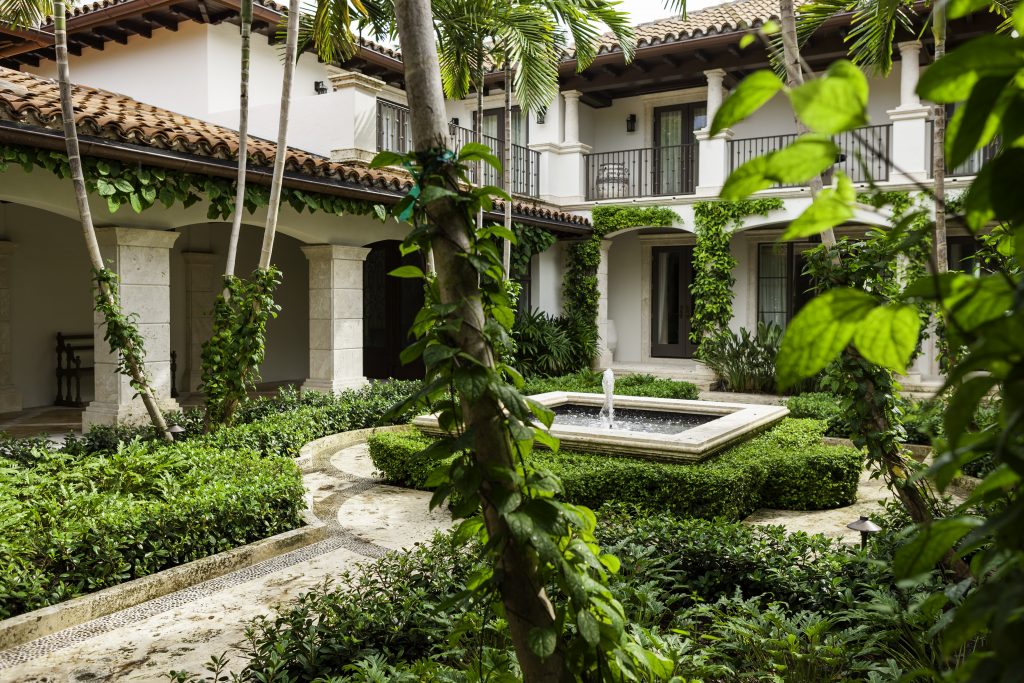 Spanish-style courtyard with a central fountain and lush greenery, surrounded by a two-story building with terracotta roof and stone columns.