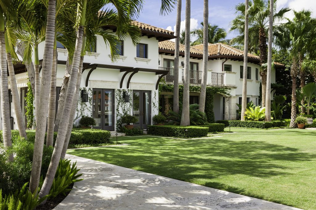 A Mediterranean-style house with terracotta roof, surrounded by palm trees and lush garden, features balconies, green lawn, and stone pathway under clear sky.
