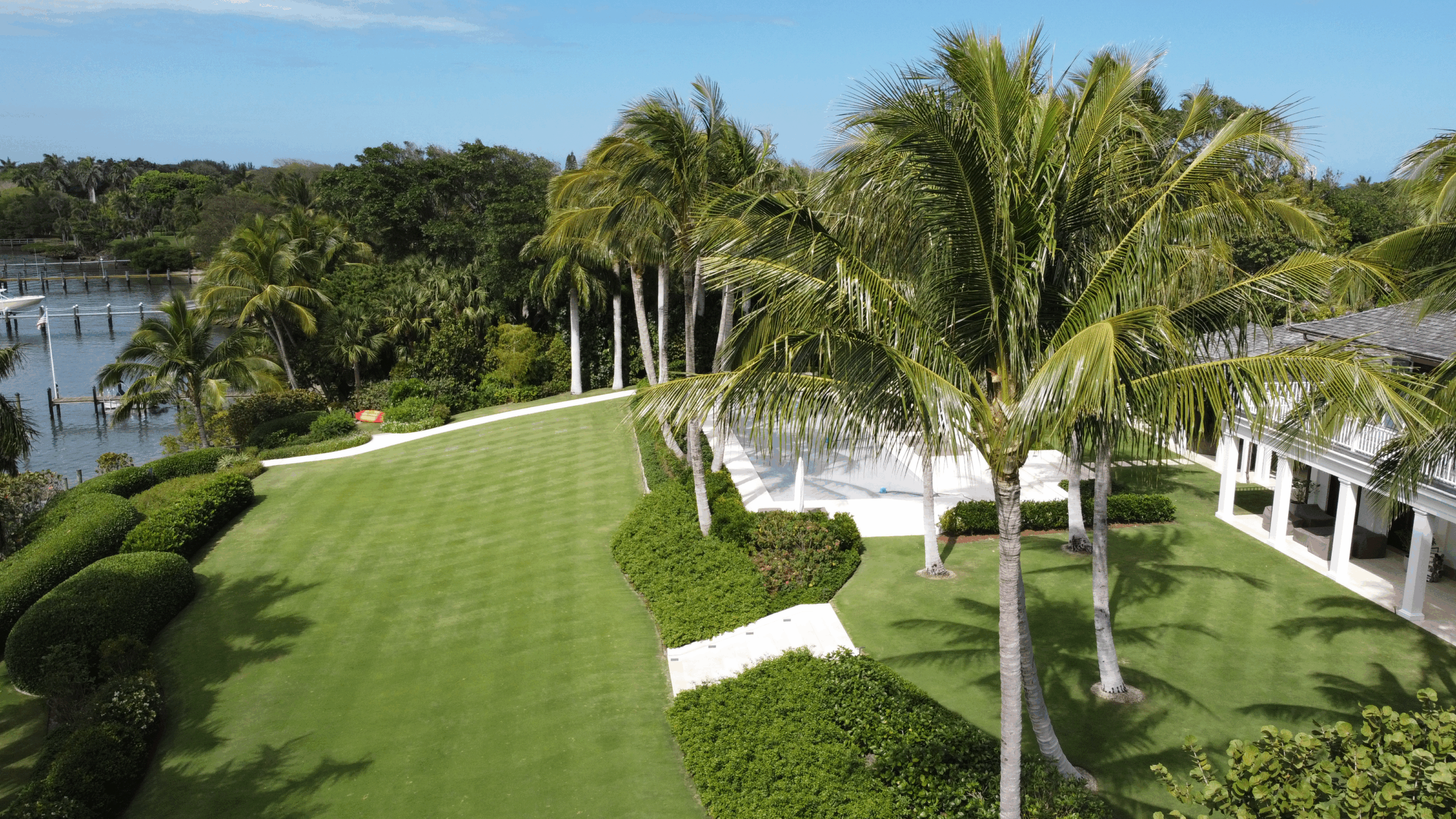 Lush tropical garden with palm trees, waterfront, and part of a large white building. Clear blue sky and manicured lawn suggest a serene setting.