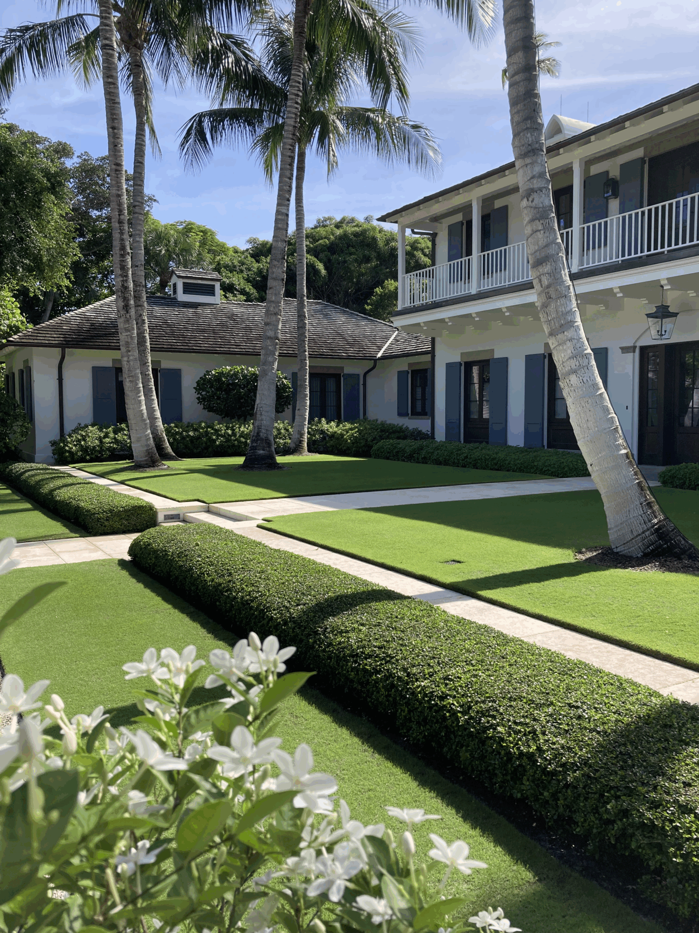 A lush garden with neatly trimmed hedges and palm trees surrounds a white house featuring blue shutters and a wraparound balcony.