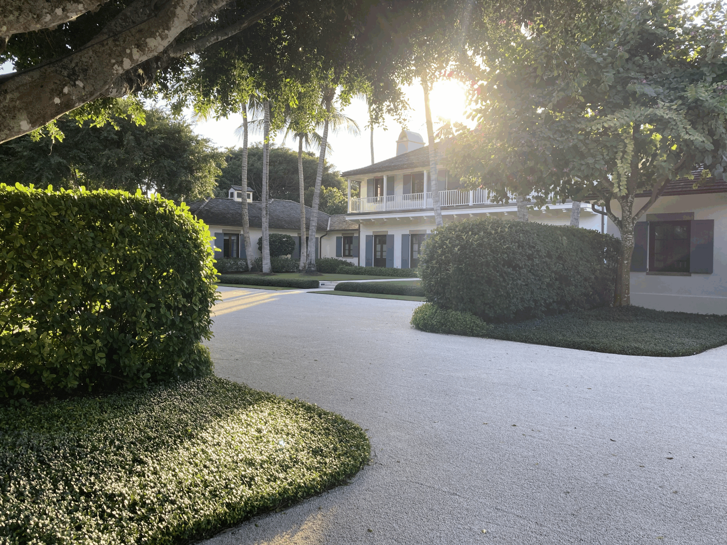 A sunlit, serene property with lush greenery, a two-story house, and palm trees. No recognizable landmarks or historical buildings present.
