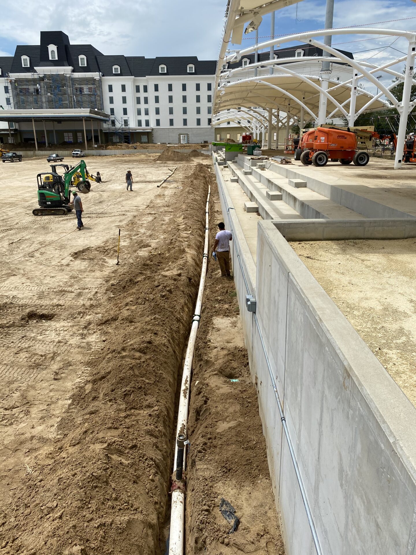 Three people work at a construction site with vehicles, near a large building resembling a hotel and covered pavilion structure.