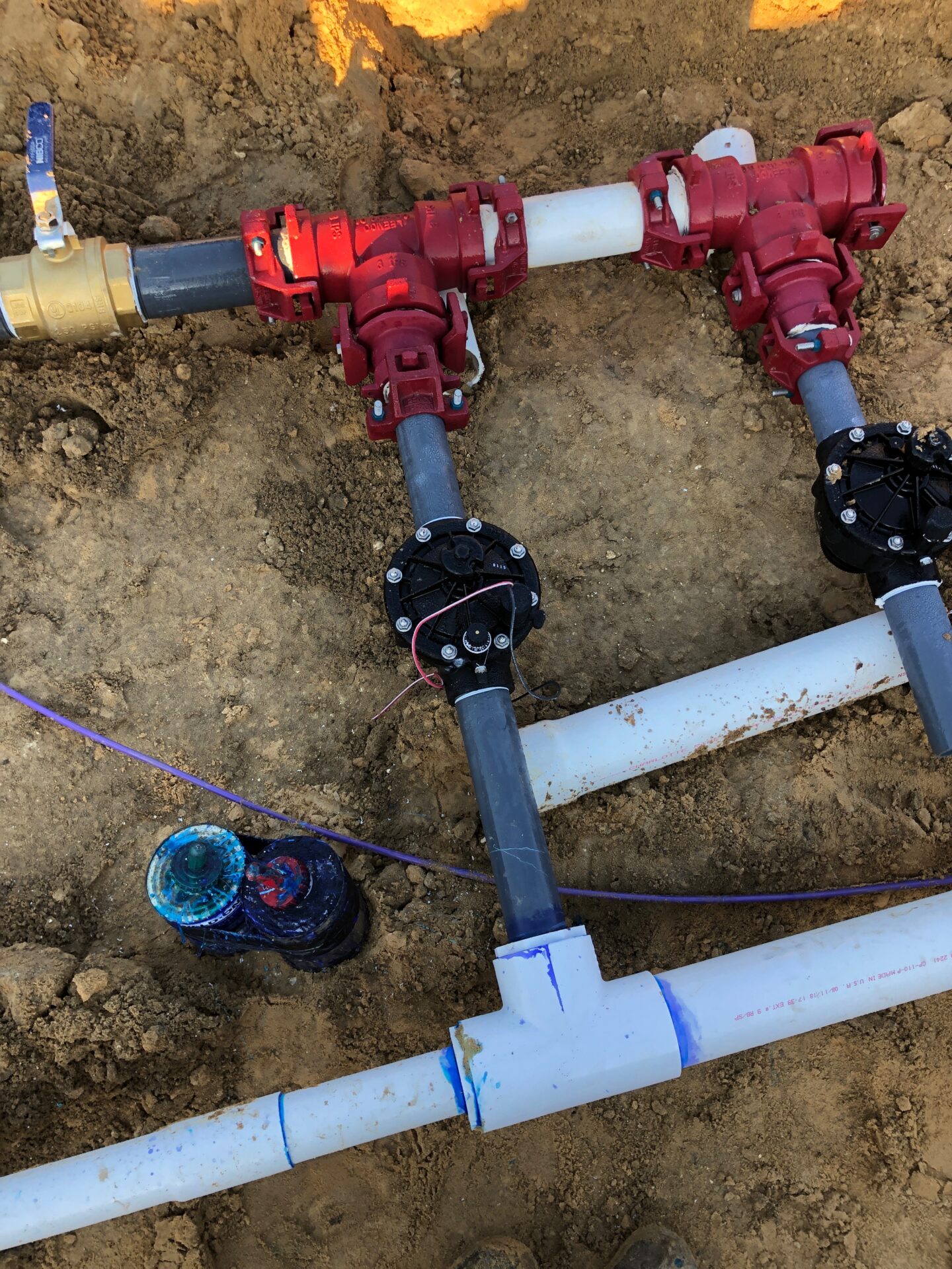 Pipes and valves interconnected in a trench, featuring red connectors and blue markings on a sandy surface, demonstrating construction or plumbing work.