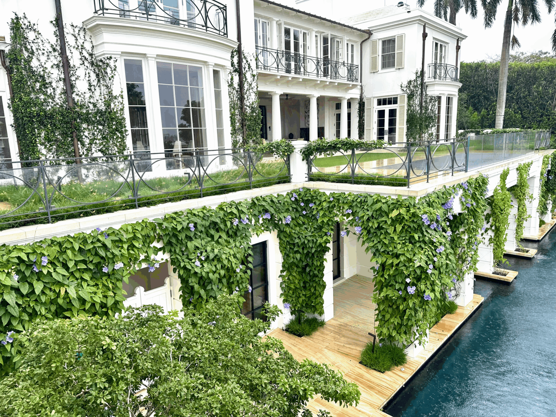 Elegant white building with large windows, adorned with lush green vines and purple flowers, beside a tranquil pool, surrounded by greenery.