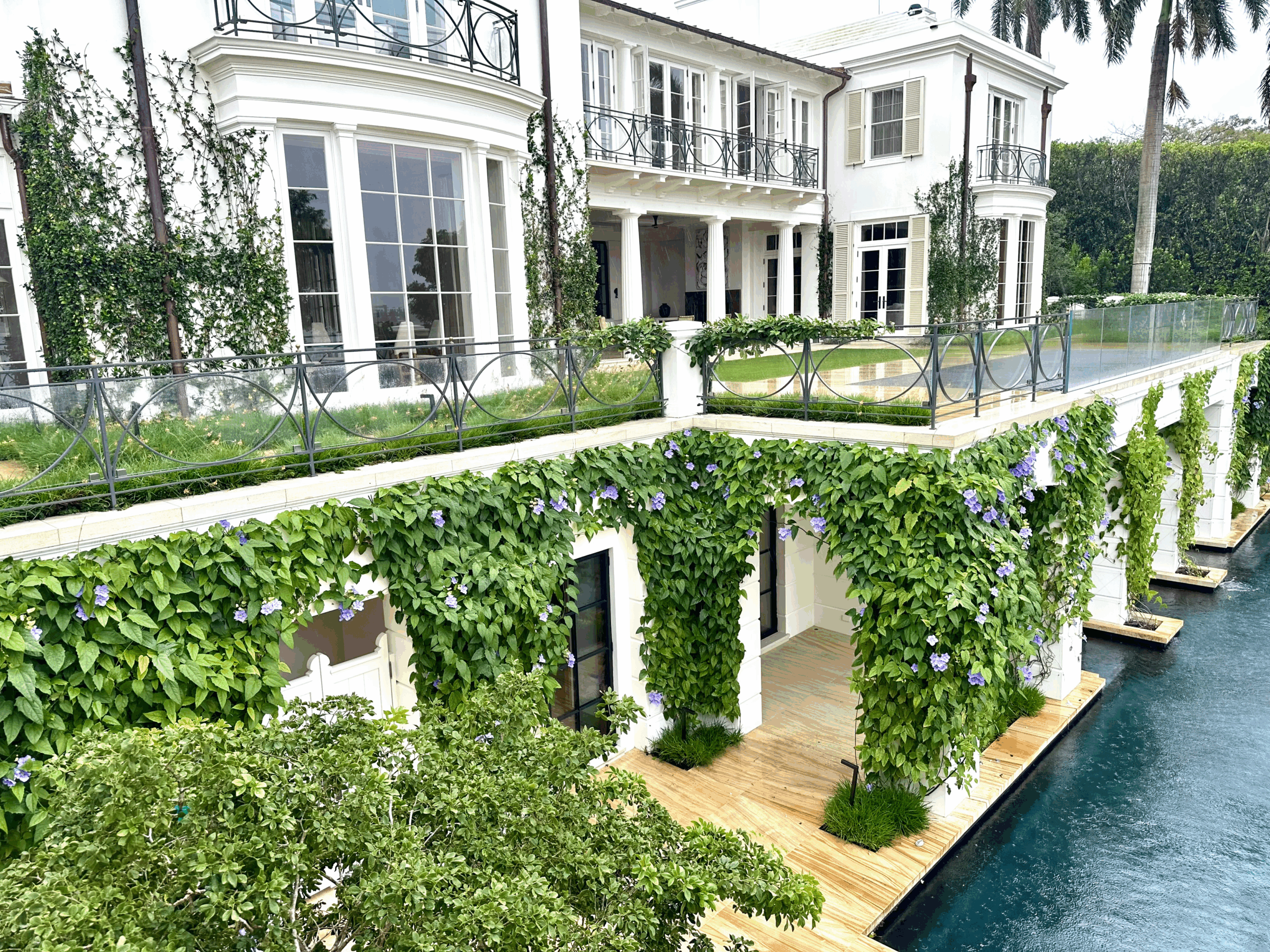 Elegant white building with large windows, adorned with lush green vines and purple flowers, beside a tranquil pool, surrounded by greenery.