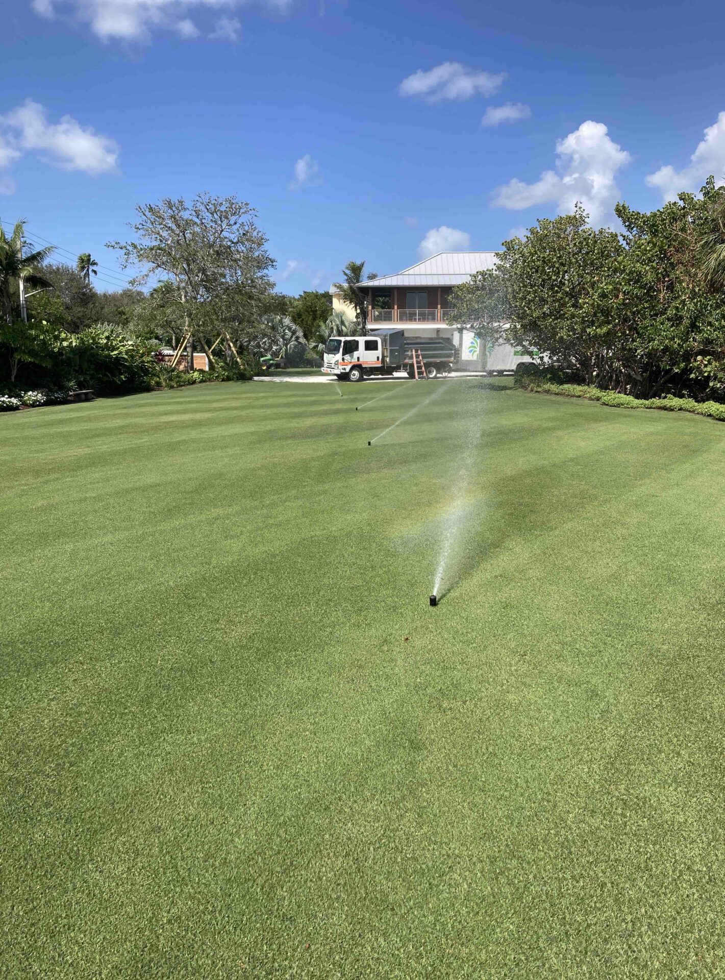 A sunny backyard scene features a well-maintained lawn with active sprinklers, a parked utility truck, and lush greenery surrounding a house.
