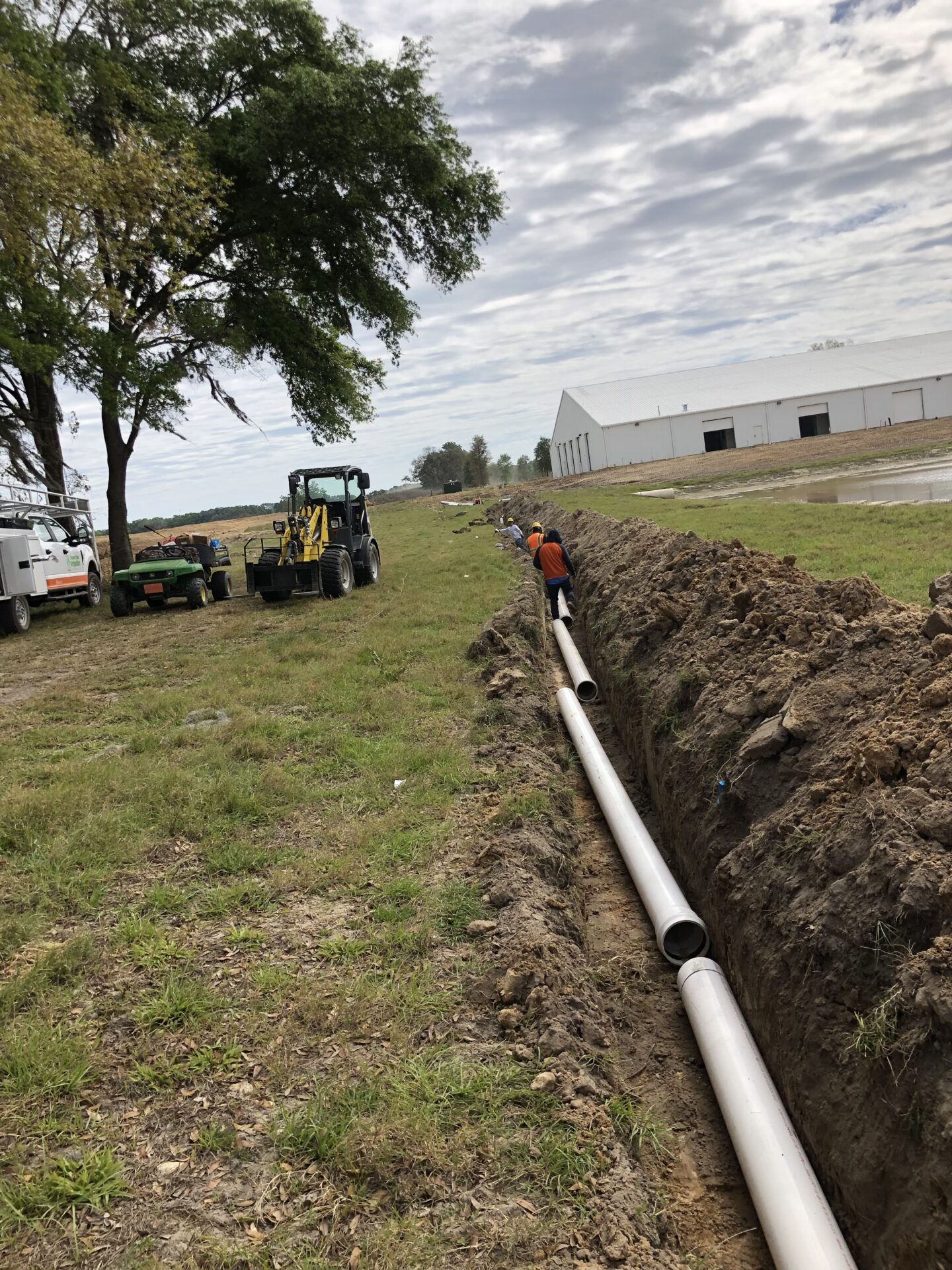 A person works in a trench with pipes, surrounded by construction vehicles, trees, and a large white industrial building under a cloudy sky.
