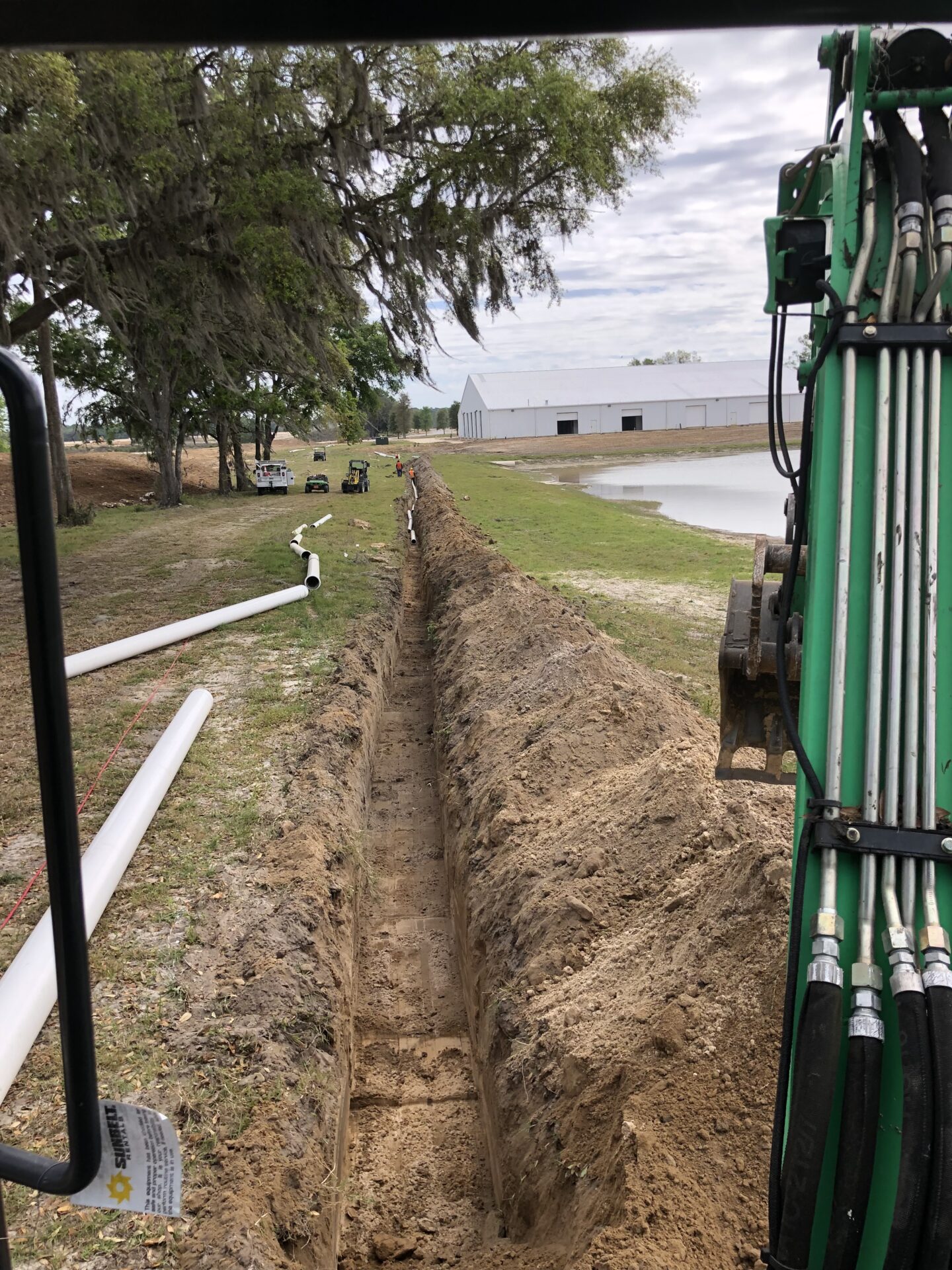An excavator digs a long trench near a pond, while pipes are laid out alongside. People and vehicles are visible under large trees.
