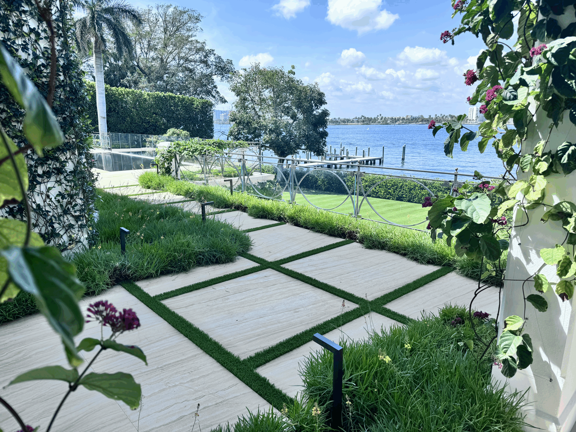 A beautifully landscaped garden with geometric paths overlooks a serene waterfront, framed by lush greenery and flowering vines under a clear blue sky.