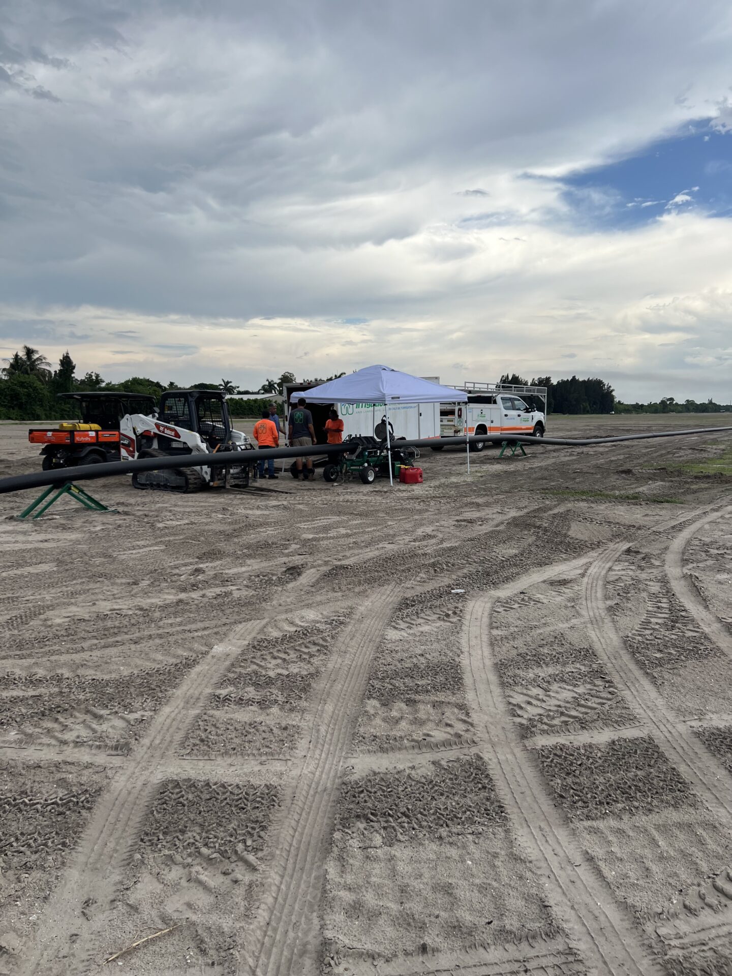 A construction site with vehicles, a tent, and people working under a cloudy sky. Tire tracks cover the sandy ground.