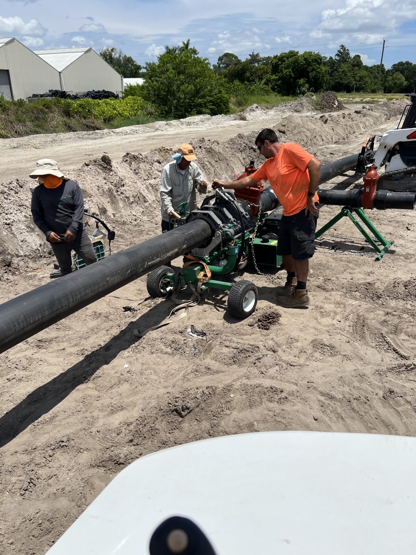 Three people work on a construction site, managing a large black pipe with machinery. The surroundings include dirt, greenery, and industrial buildings.