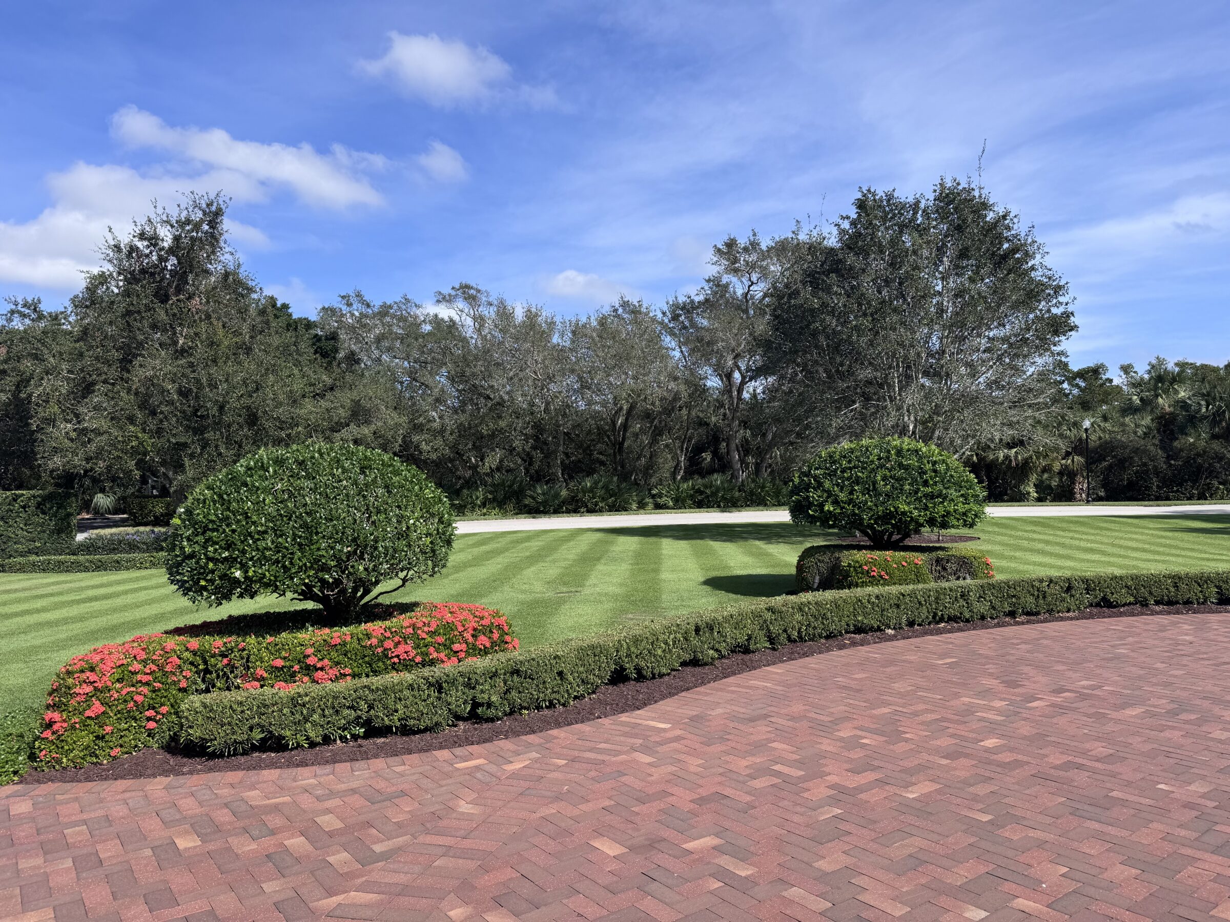 Manicured lawn with striped grass patterns, circular shrubs, and vibrant flowers, under a clear blue sky. Trees line the background, creating a serene atmosphere.