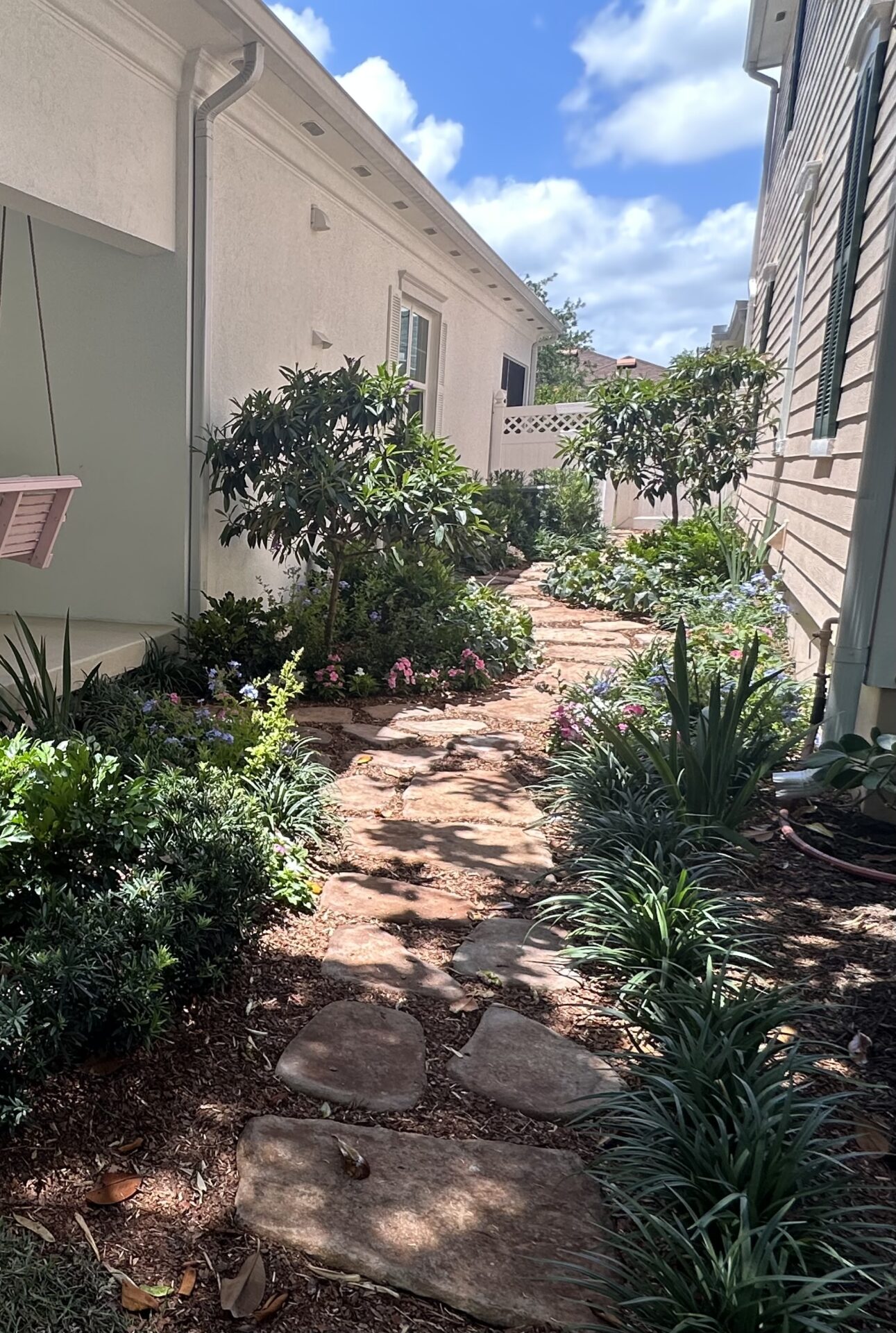 A stone pathway lined with lush greenery and colorful flowers between two buildings under a partly cloudy sky. Peaceful and inviting garden scene.