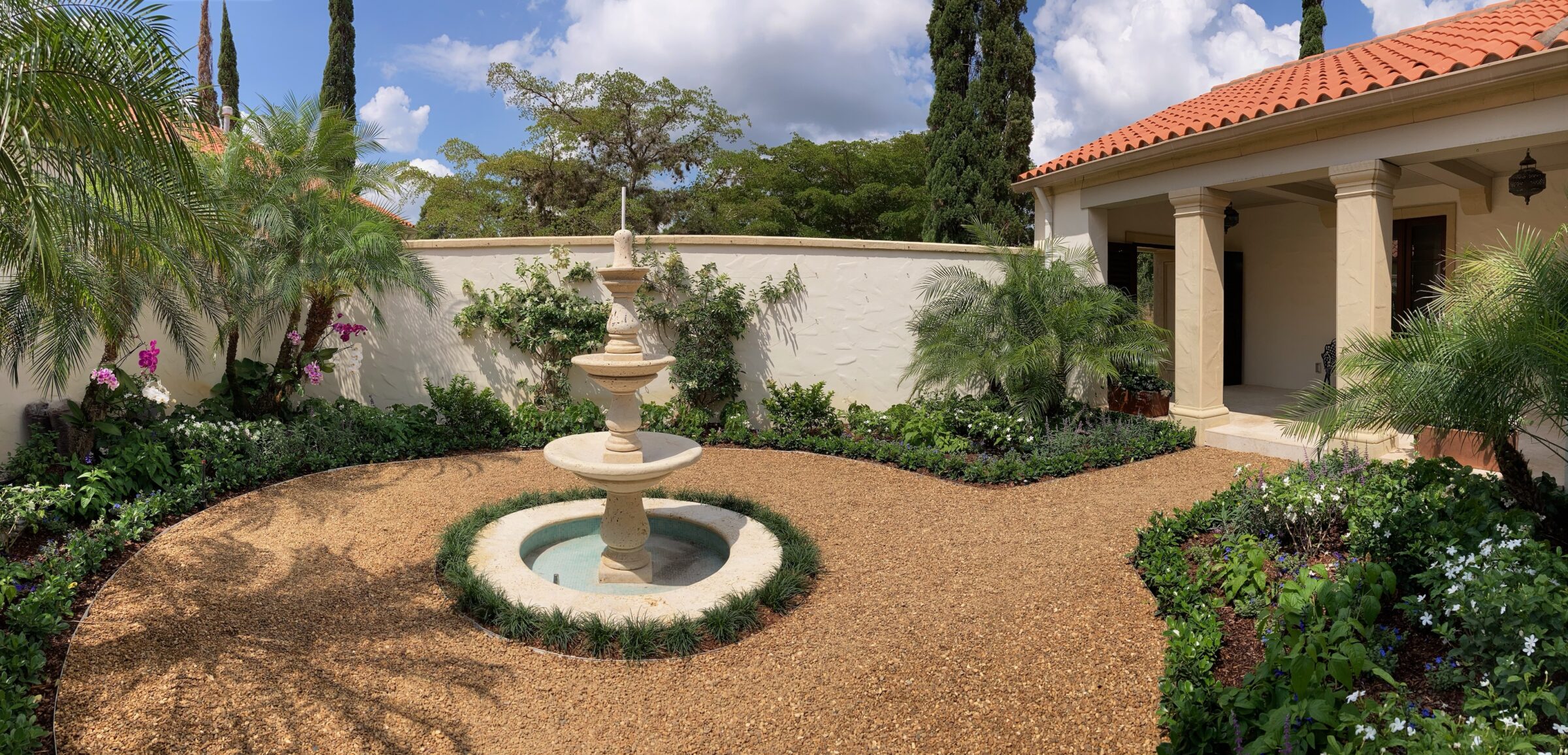 A Mediterranean-style courtyard with a stone fountain, surrounded by lush greenery, palm trees, and a building with terracotta roof tiles.