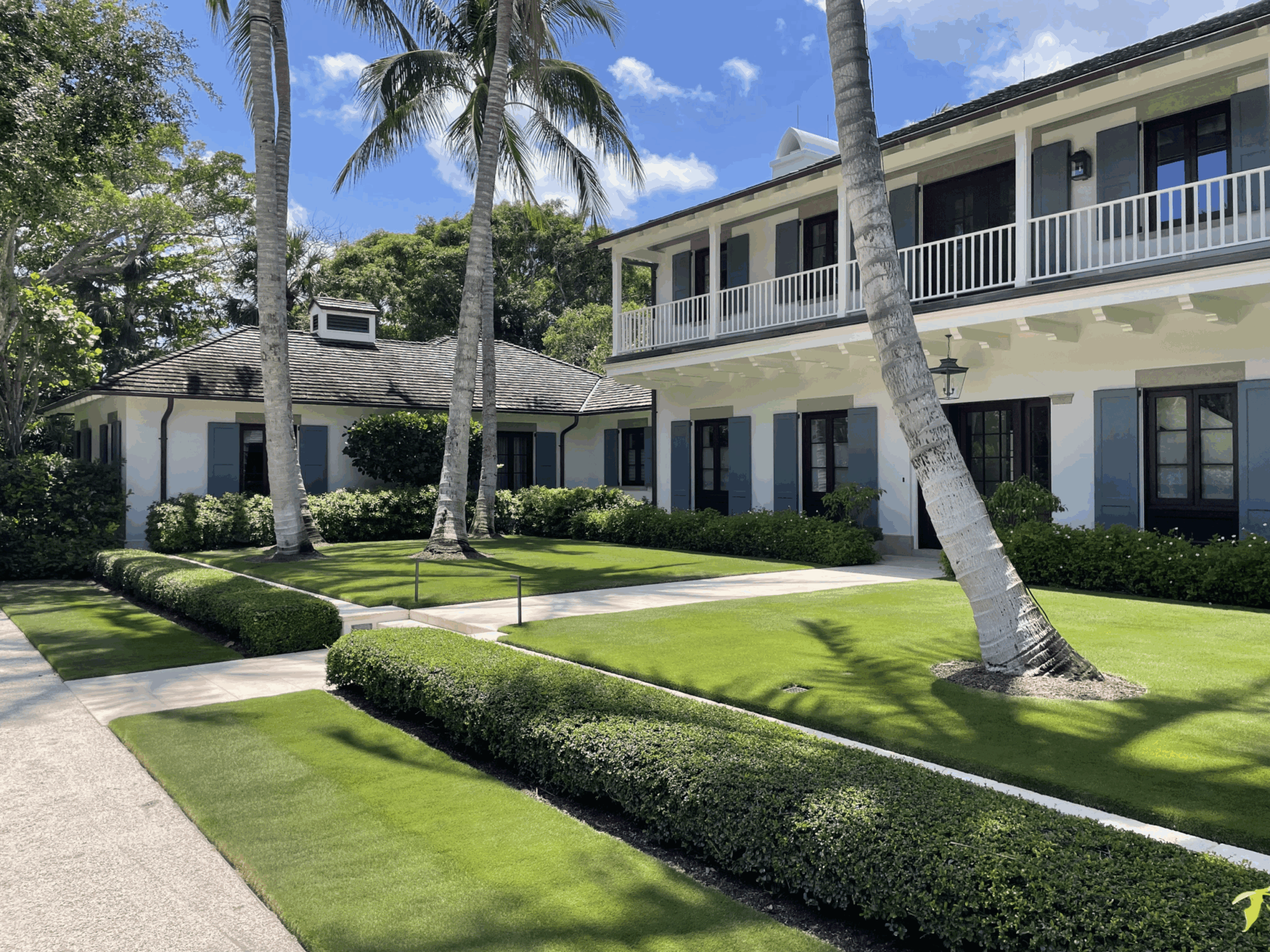 A charming, colonial-style building with white walls and blue shutters is surrounded by lush green grass, palm trees, and hedges under a clear sky.