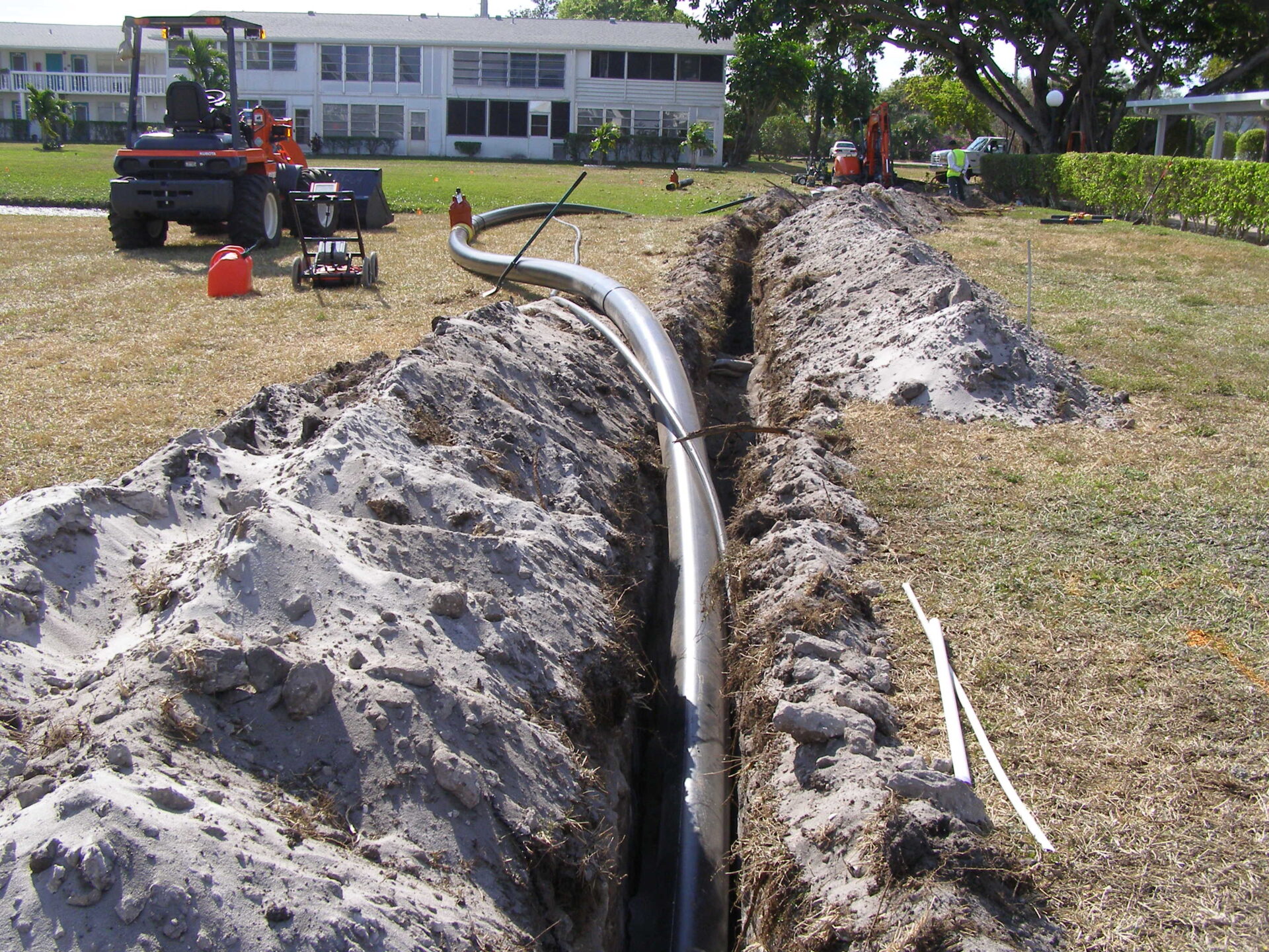 A trench with a pipe, machinery, and workers on a grassy area near residential buildings under construction in a sunny outdoor setting.