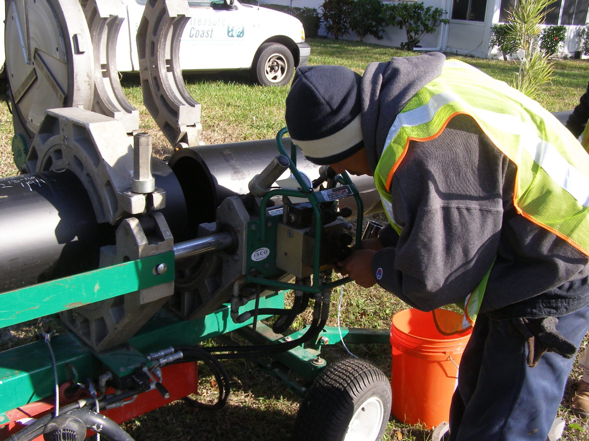 A person in a safety vest operates a pipe fusion machine outdoors near a vehicle marked 