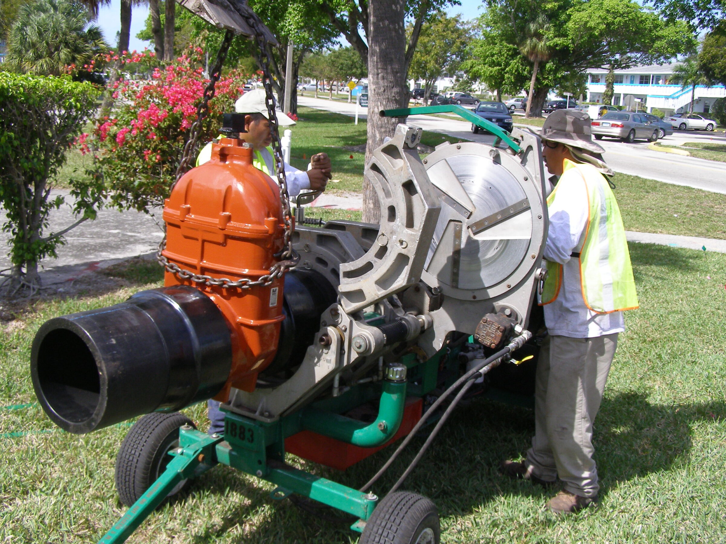 Two people operate a large, industrial pipe fusion machine on a grassy area near a road, surrounded by trees and bushes.