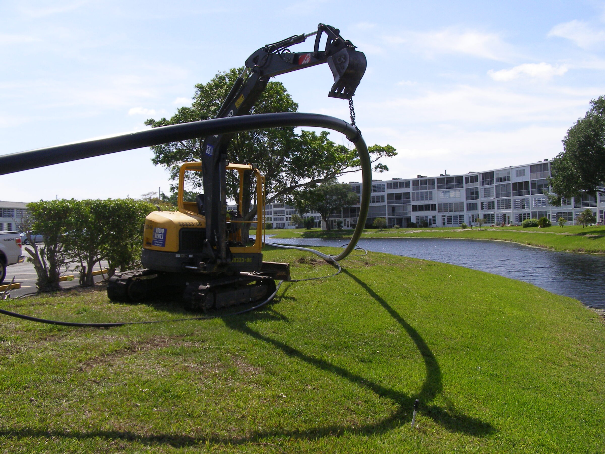 A construction vehicle manipulates a long hose on grassy terrain near a canal, with residential buildings visible in the background.