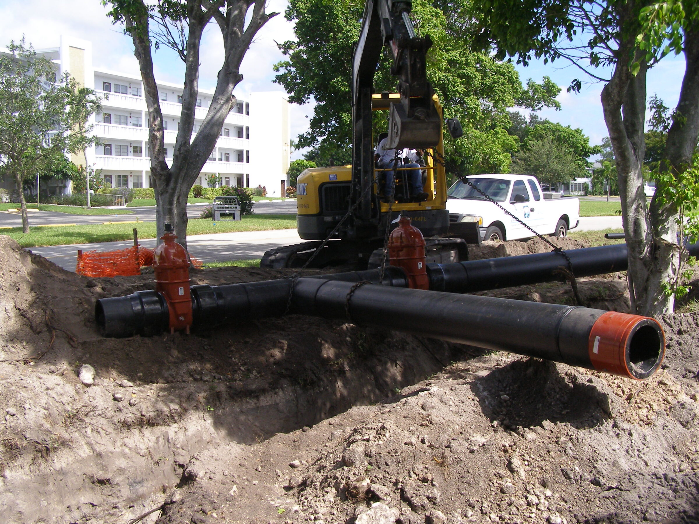 A construction site with heavy machinery installing large black pipes near a residential building, surrounded by trees and parked vehicles.