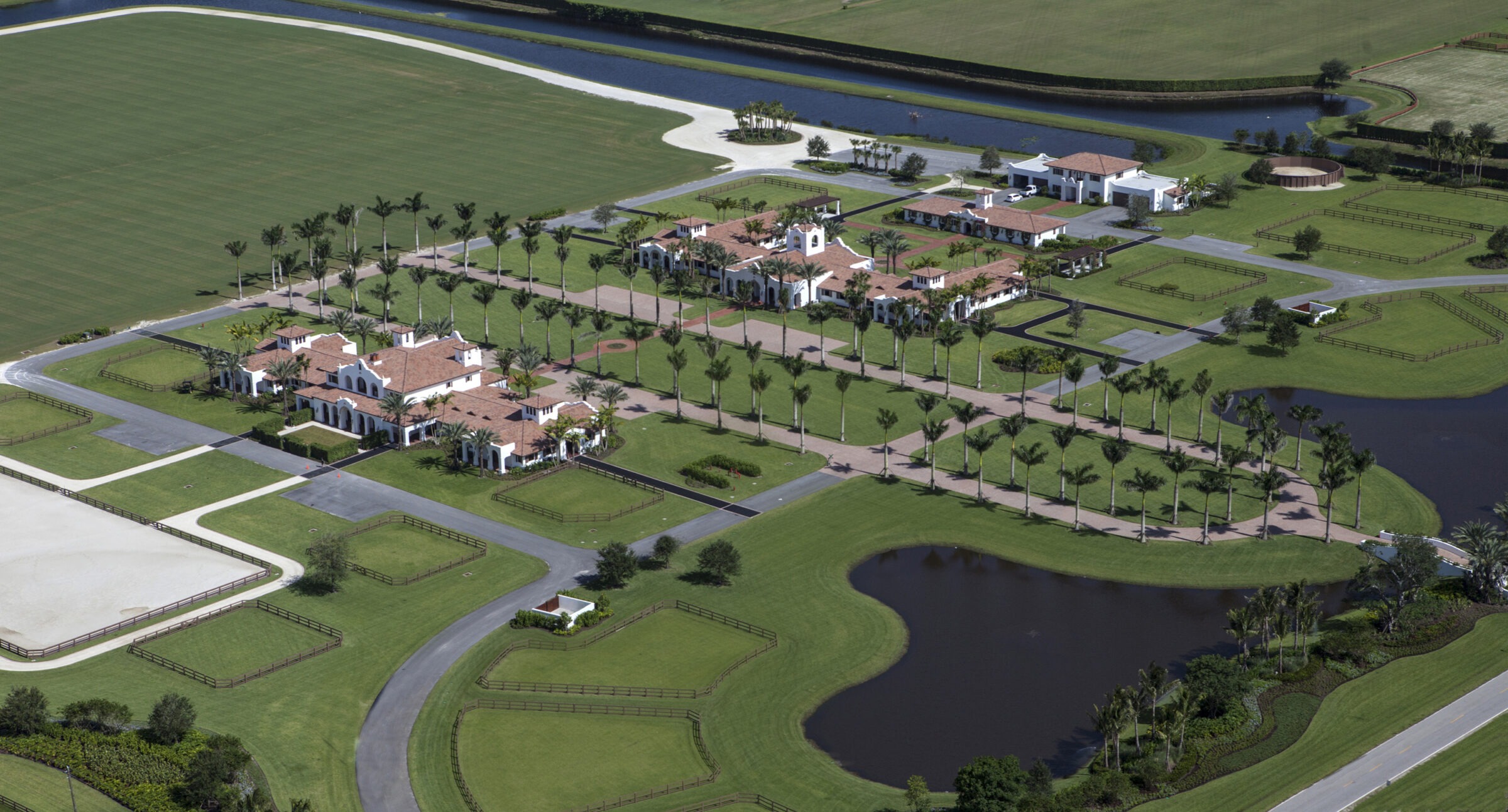 Aerial view of a luxurious estate with multiple buildings, palm-lined driveways, surrounding water features, and expansive green lawns. No landmarks visible.