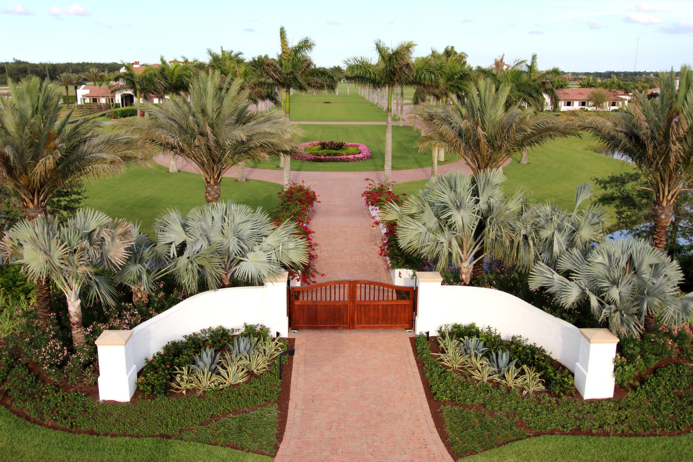 A beautifully landscaped garden path with palm trees, flowers, and a wooden gate leading to a large grassy area with distant structures.