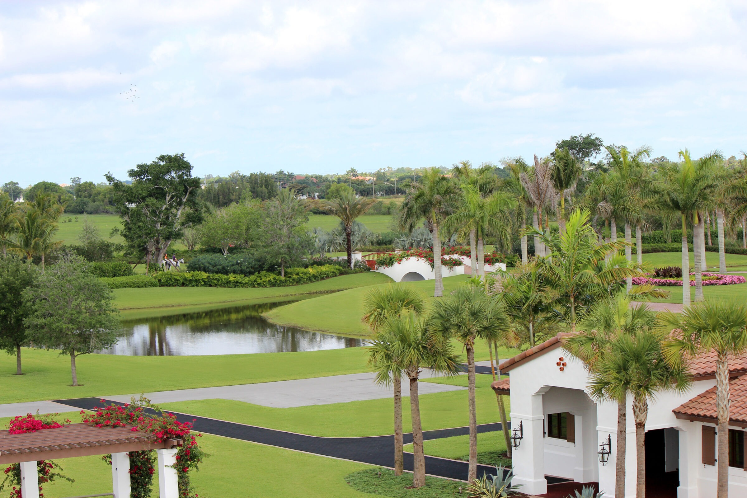 A lush golf course landscape featuring a white building, palm trees, a small pond, and a bridge under a bright, partly cloudy sky.