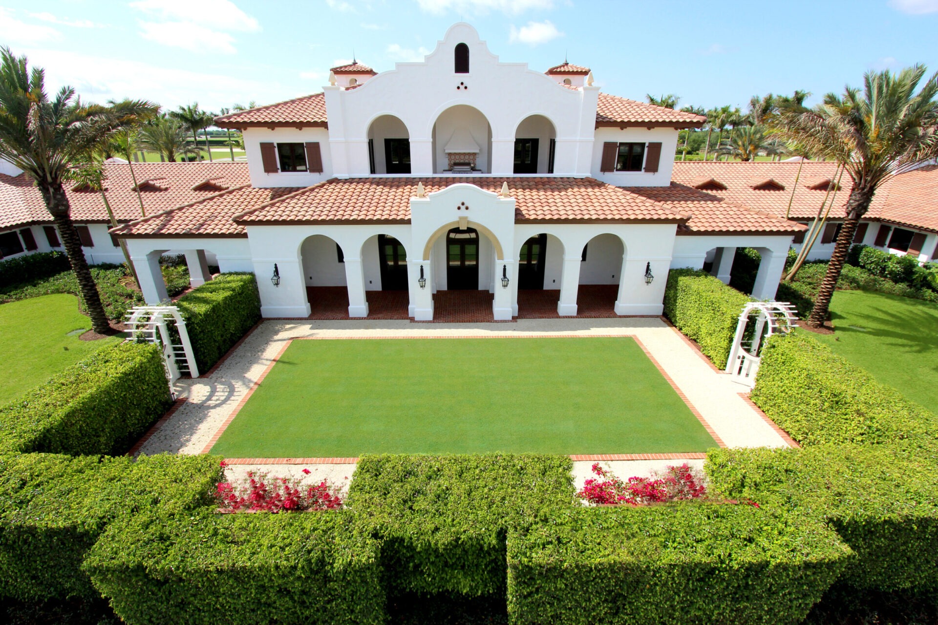 Spanish-style villa with white walls, red roof tiles, arched entrances, surrounded by lush greenery, symmetrical garden, and palm trees under a clear blue sky.