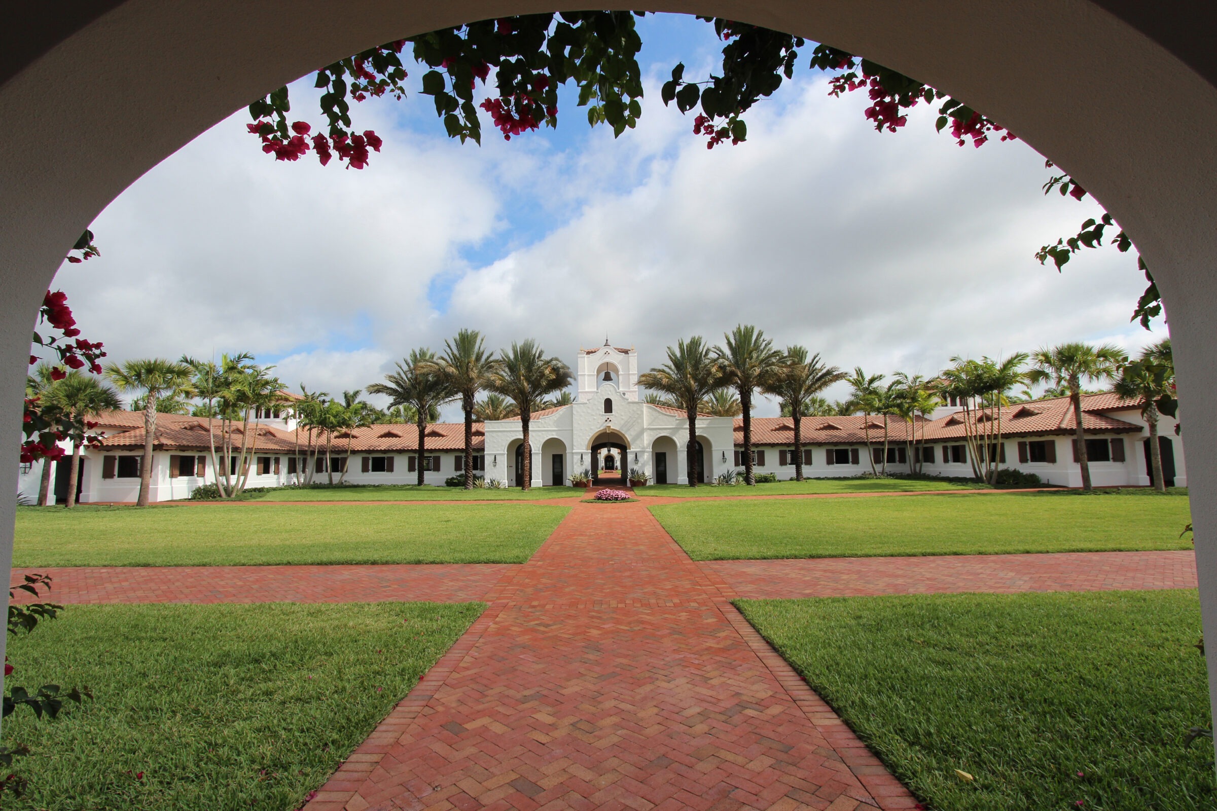 Arched view of lush courtyard with red brick path, palm trees, and a historic white stucco building under a partly cloudy sky.