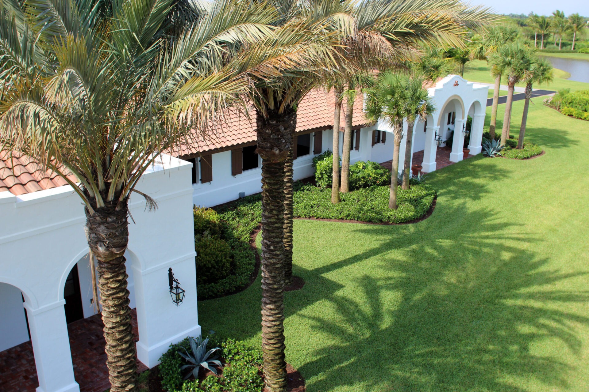 A white building with arches and a red-tiled roof, surrounded by palm trees and lush green lawn, near a small pond.