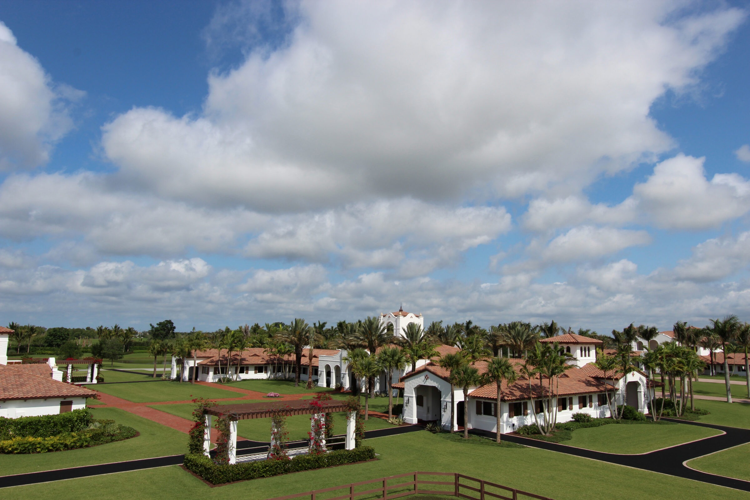 Lush landscape featuring white villas with terracotta roofs, palm trees, and a pergola under a cloudy blue sky. Peaceful and idyllic setting.