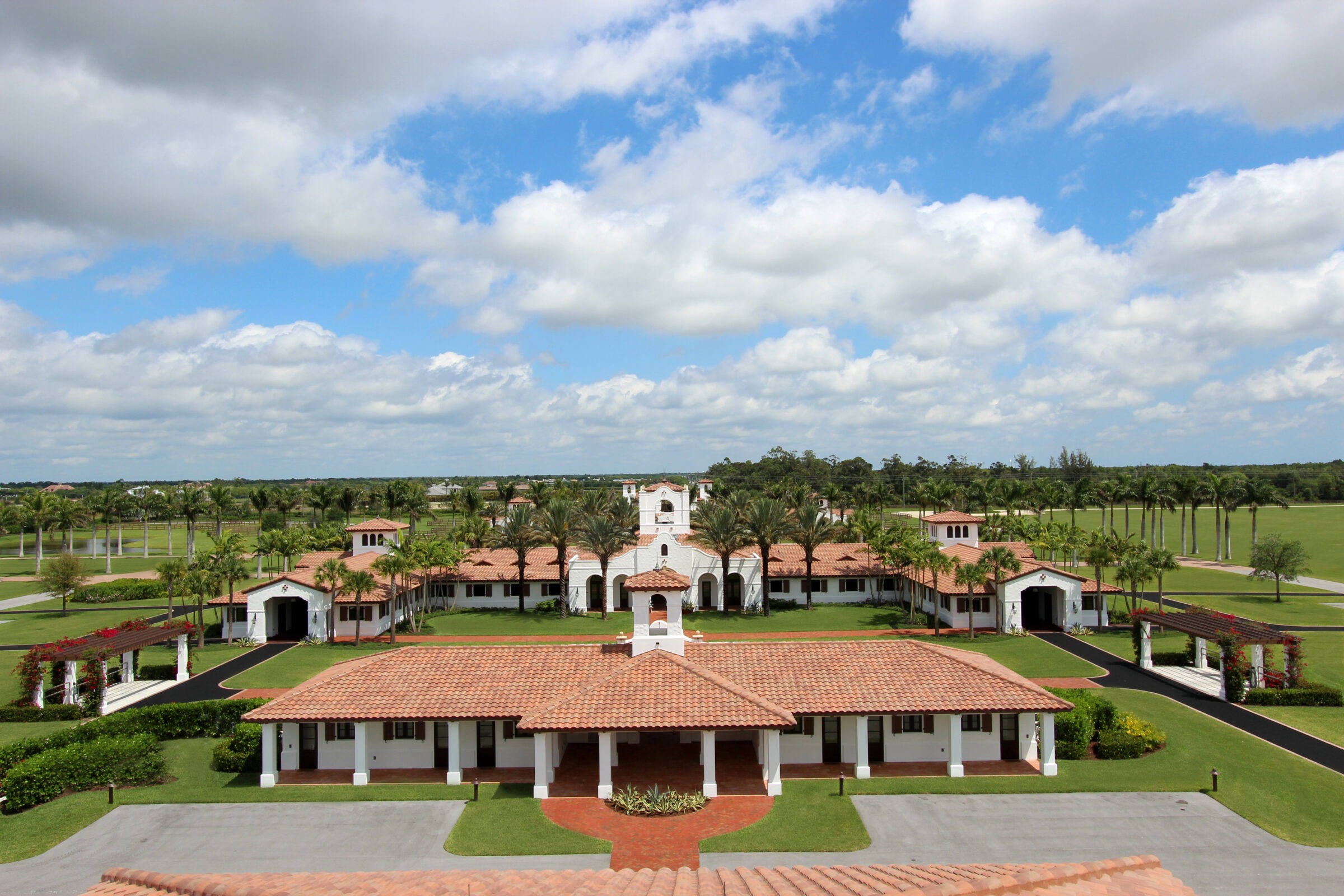 Sunny landscape featuring a large estate with red-tiled roofs, palm trees, and expansive lawns under a partly cloudy sky.