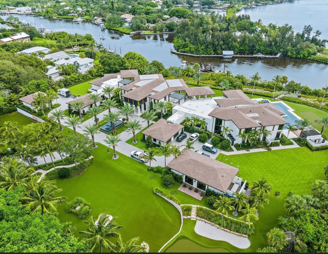 Aerial view of a luxurious estate with large buildings, green lawns, palm trees, pool, and waterfront access, surrounded by lush vegetation and a river.