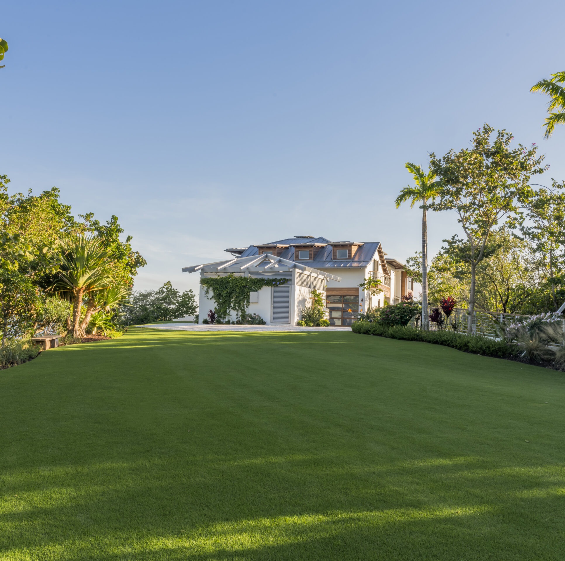 A modern house with a large lawn, surrounded by trees and greenery under a clear blue sky. Sunlight enhances the vibrant landscape.