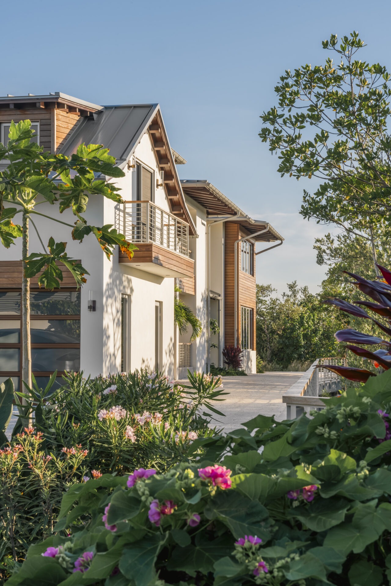Modern house with wooden accents and balconies, surrounded by lush greenery and colorful flowers under a clear blue sky. Quiet, serene atmosphere.