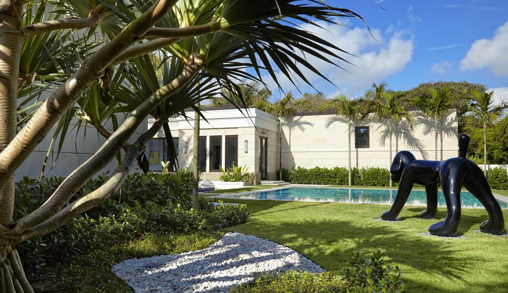 Modern backyard with a large black dog sculpture, pool, palm trees, and a person near a contemporary building on a sunny day.