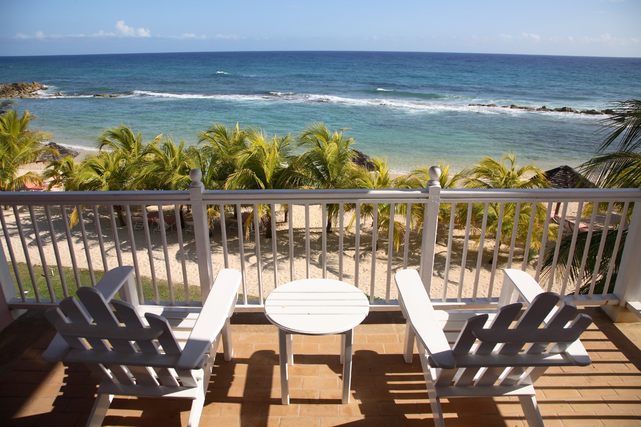 White chairs and table on a balcony overlook a sandy beach with palm trees and turquoise ocean, under a clear blue sky.