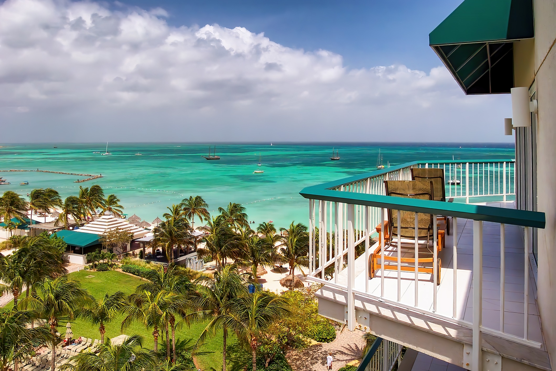 Balcony view overlooking a turquoise ocean, palm trees, and sandy beach. Several boats are anchored in the distance under a partly cloudy sky.