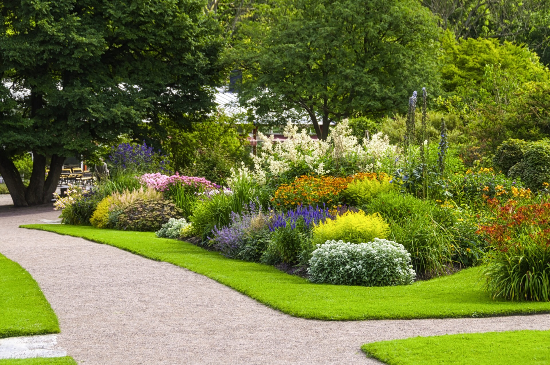 Lush garden with colorful flowers and green paths surrounded by trees. A person sits in the distance, partially hidden by foliage.