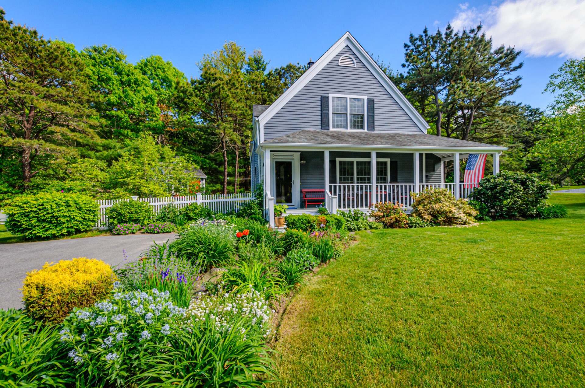 A charming gray house with a porch and American flag, surrounded by lush gardens and vibrant greenery under a clear blue sky.