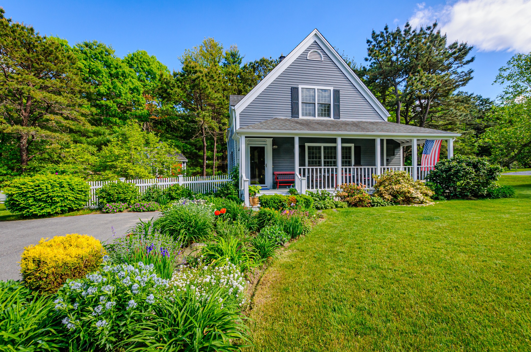 A charming gray house with a porch and American flag, surrounded by lush gardens and vibrant greenery under a clear blue sky.