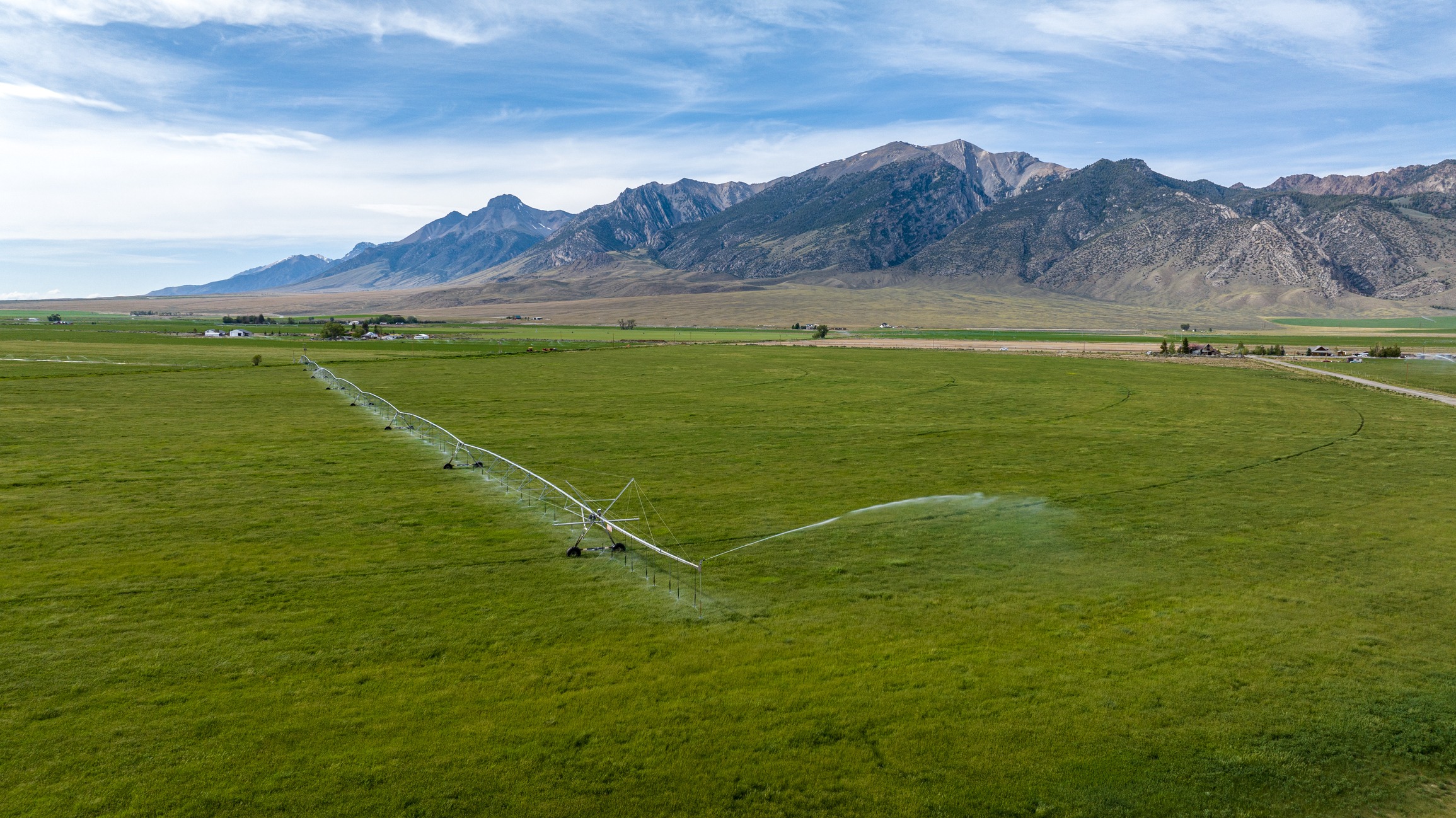 Lush green field with a central irrigation system, framed by majestic mountains under a vast blue sky, creating a serene agricultural landscape.