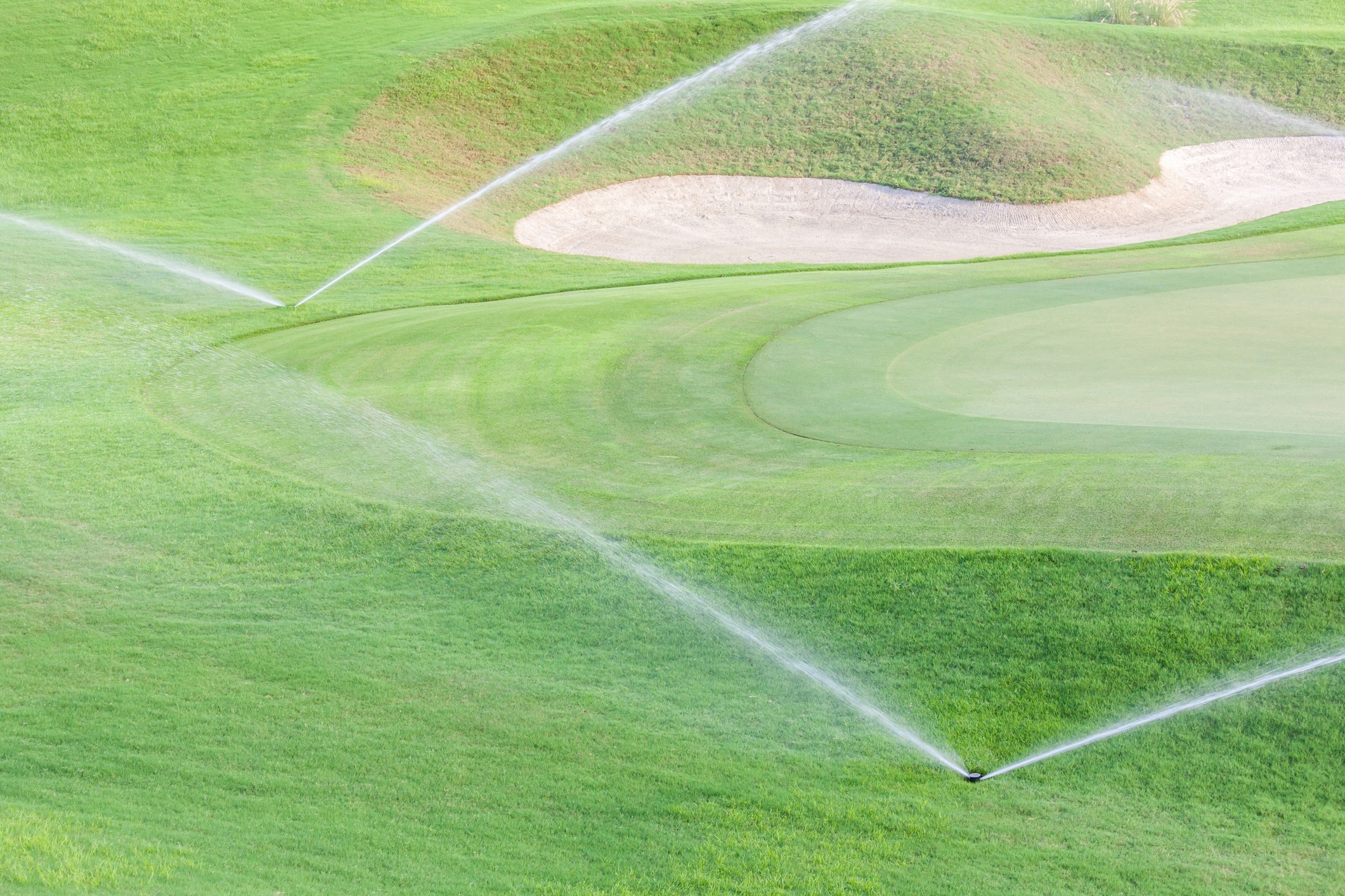 Lush golf course with sprinklers watering vibrant green grass and sand trap, creating a serene and well-maintained landscape, viewed under soft light.
