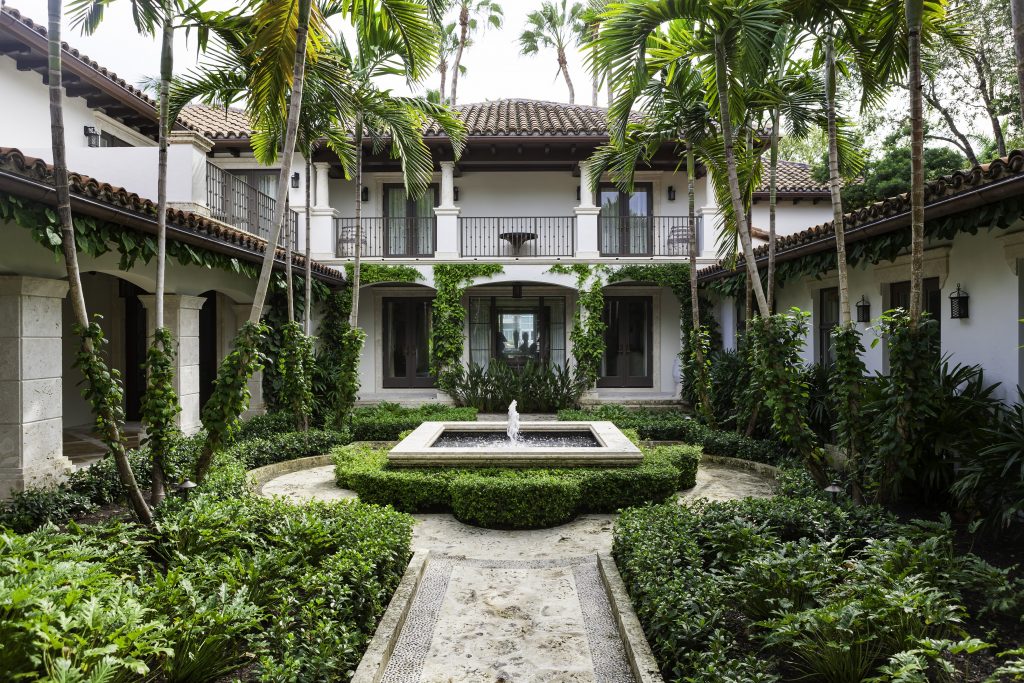 Courtyard with a central fountain, surrounded by lush greenery and palm trees, within a two-story Spanish-style building featuring tiled roofs and balconies.