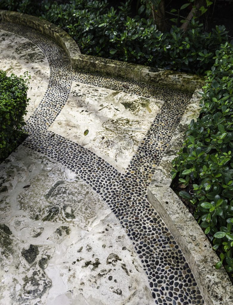 Stone pathway with intricate pebble mosaic design, bordered by lush green shrubbery, creating a serene garden atmosphere with detailed natural textures.