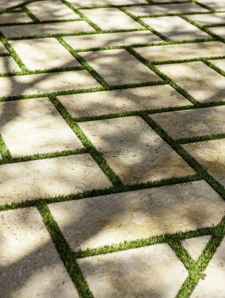 Sunlit stone tiles with grass borders create a geometric pattern on the ground, casting soft shadows from nearby trees, showcasing natural texture.