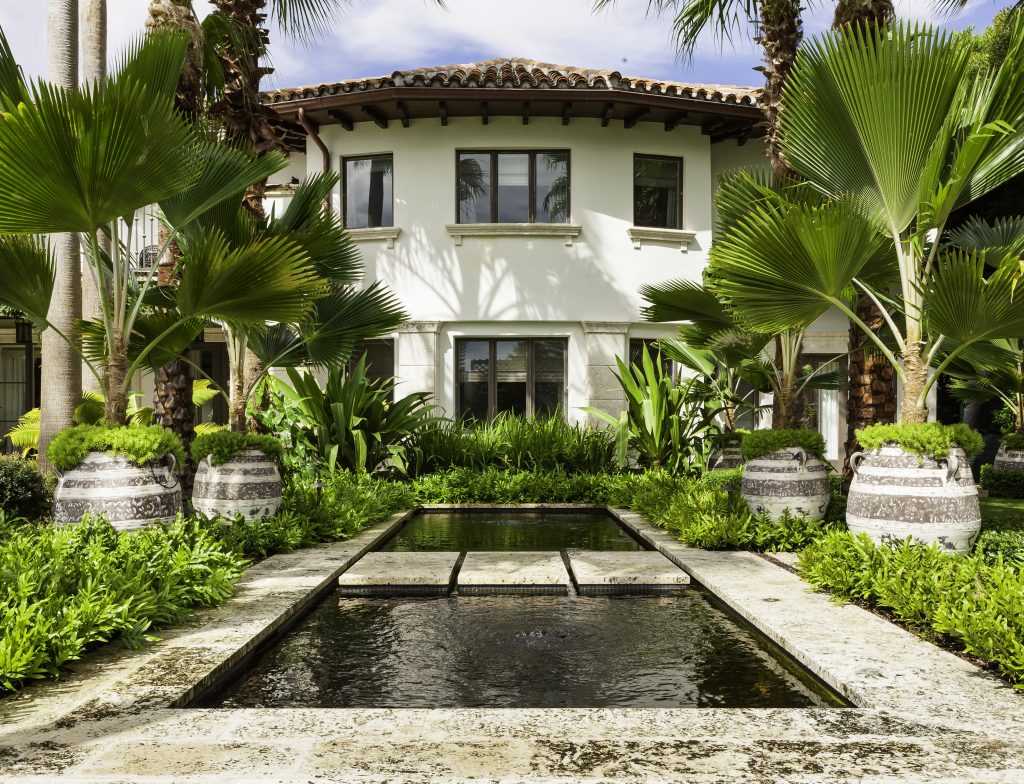 Lush garden with palm trees, large decorative pots, and a serene rectangular water feature, set before a two-story Mediterranean-style villa.