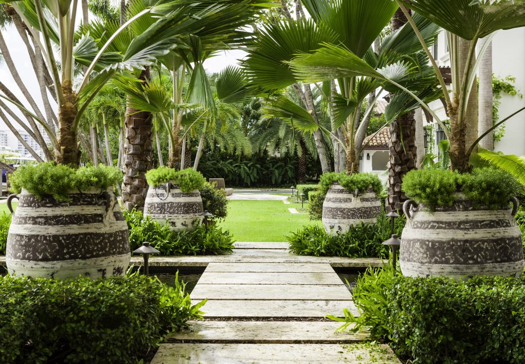 A lush garden path with large potted plants, palm trees, and a tiled roof building in the background, creating a serene atmosphere.