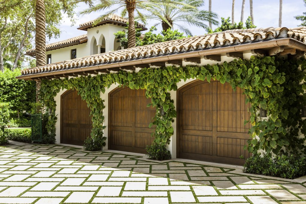 A Mediterranean-style house with a tiled roof and ivy-draped garage, surrounded by lush greenery and palm trees in a paved courtyard.